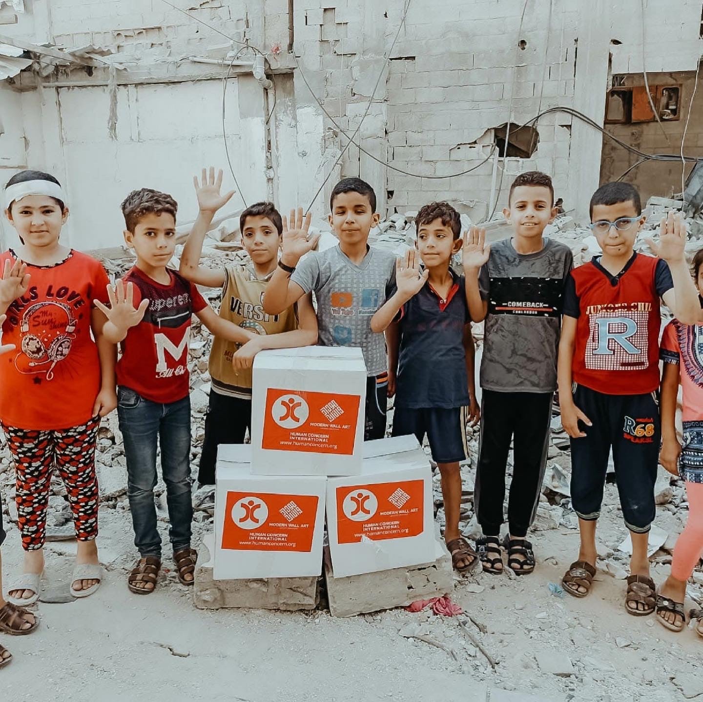 Children in a construction area, smiling and raising hands, with boxes labeled "humanitarian aid."
